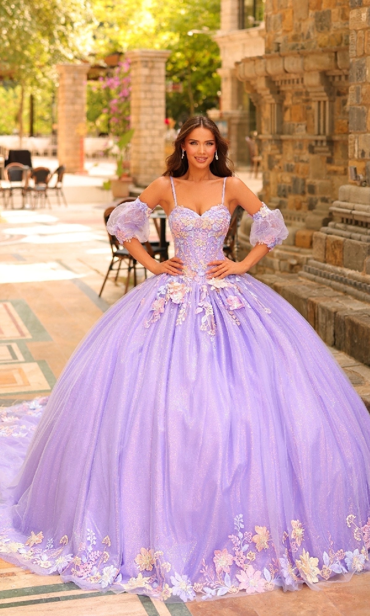 Outdoors near stone architecture and greenery, a woman poses hands on hips in the Amarra 54234 Puff-Sleeve Quinceanera Dress, featuring ornate lavender fabric with floral lace appliqués and sheer puff sleeves.